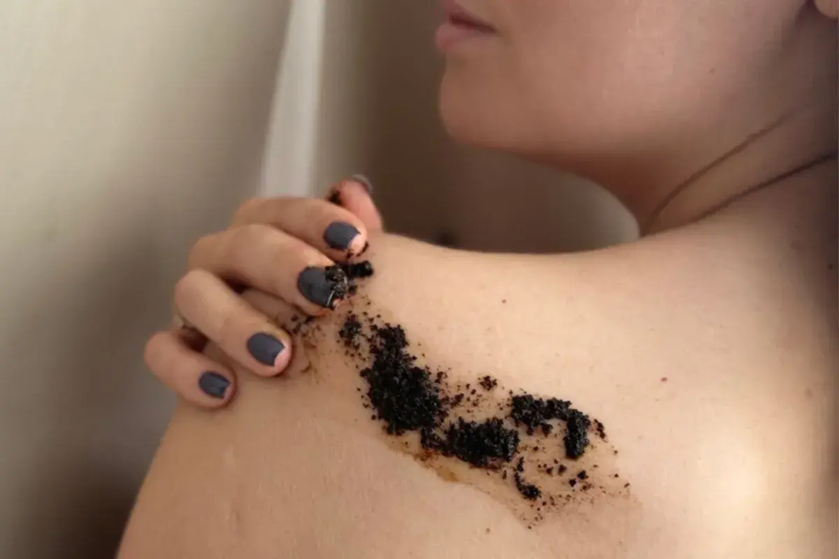 Dłoń z ciemnym lakierem do paznokci rozprowadza na ramieniu drobny, ciemny peeling z kawy.